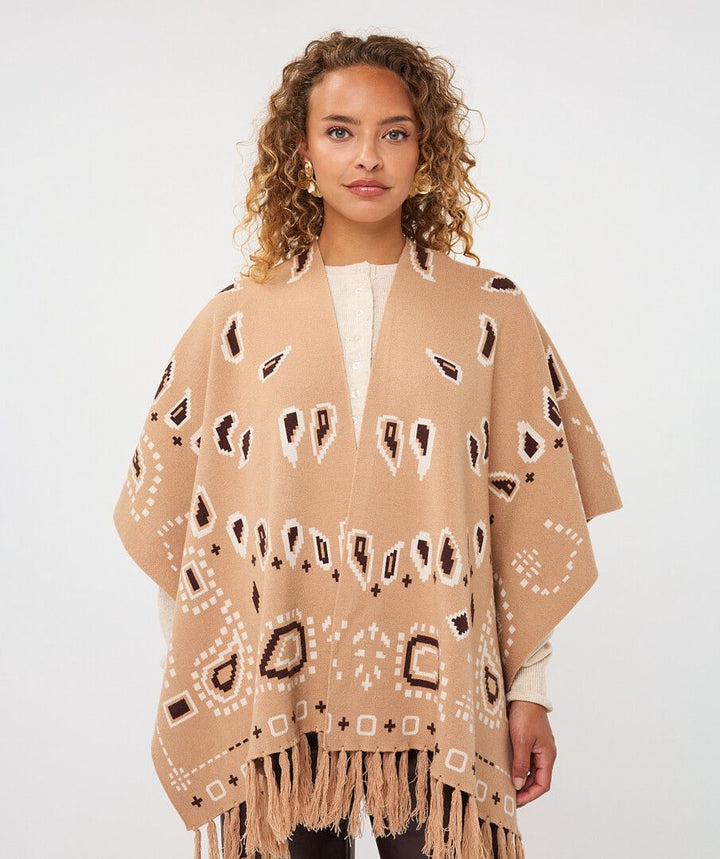 A woman wearing a beige open-front poncho with white and dark brown geometric patterns and fringe, photographed against a light gray studio background.