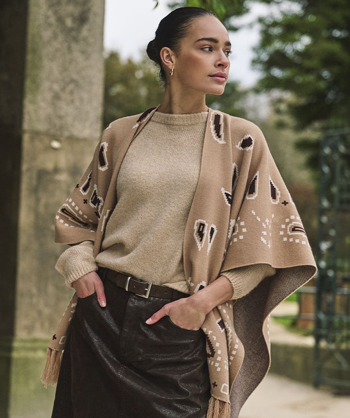 A woman stands outdoors wearing a beige knit poncho with black and white geometric patterns over a beige sweater and a dark brown skirt.