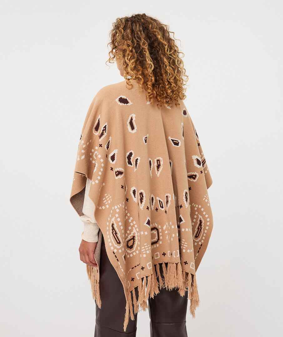 A woman shown from behind wearing a camel-colored knit poncho with white and dark brown abstract patterns and fringe, paired with dark brown pants against a light gray studio background.