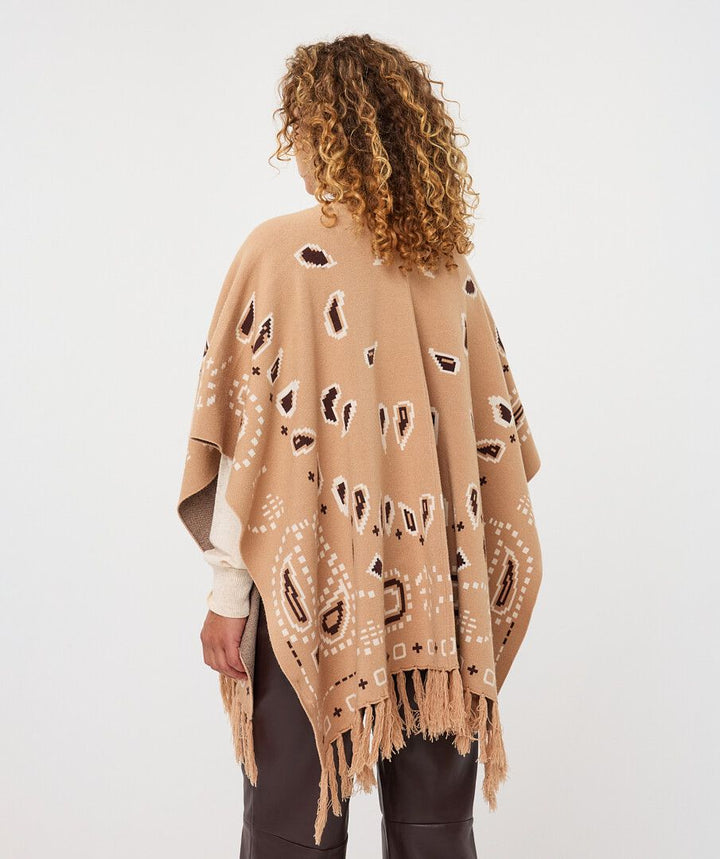 A woman shown from behind wearing a camel-colored knit poncho with white and dark brown abstract patterns and fringe, paired with dark brown pants against a light gray studio background.