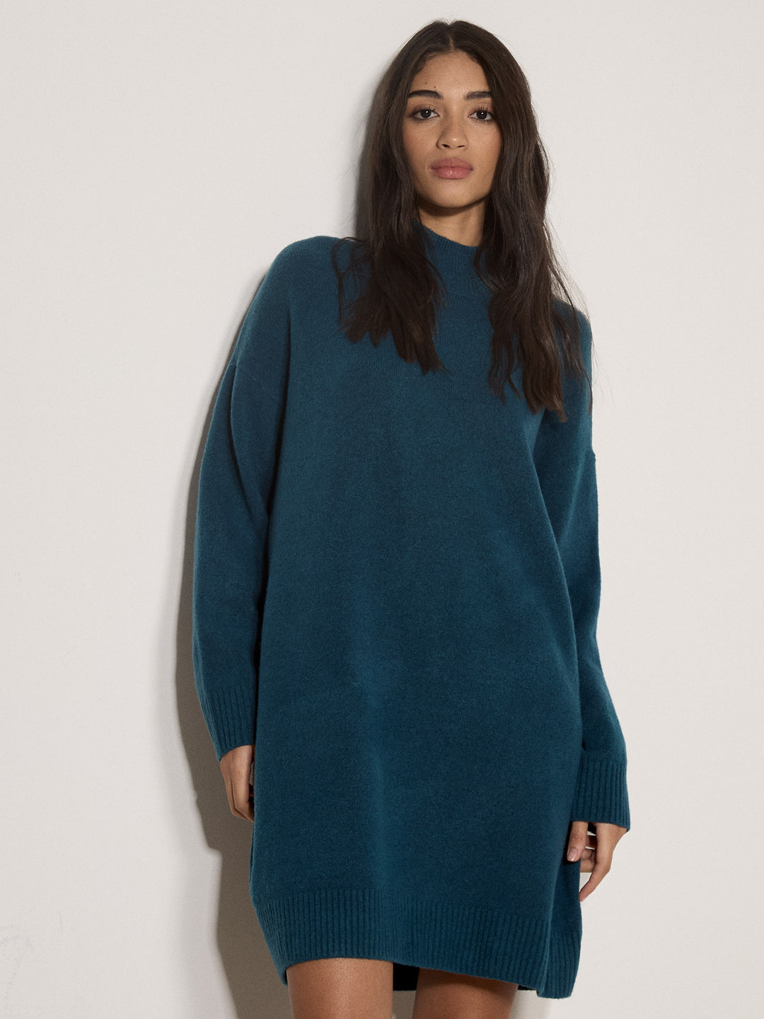 A woman modeling an oversized knit sweater dress in a deep teal blue color, standing against a plain off-white wall.
