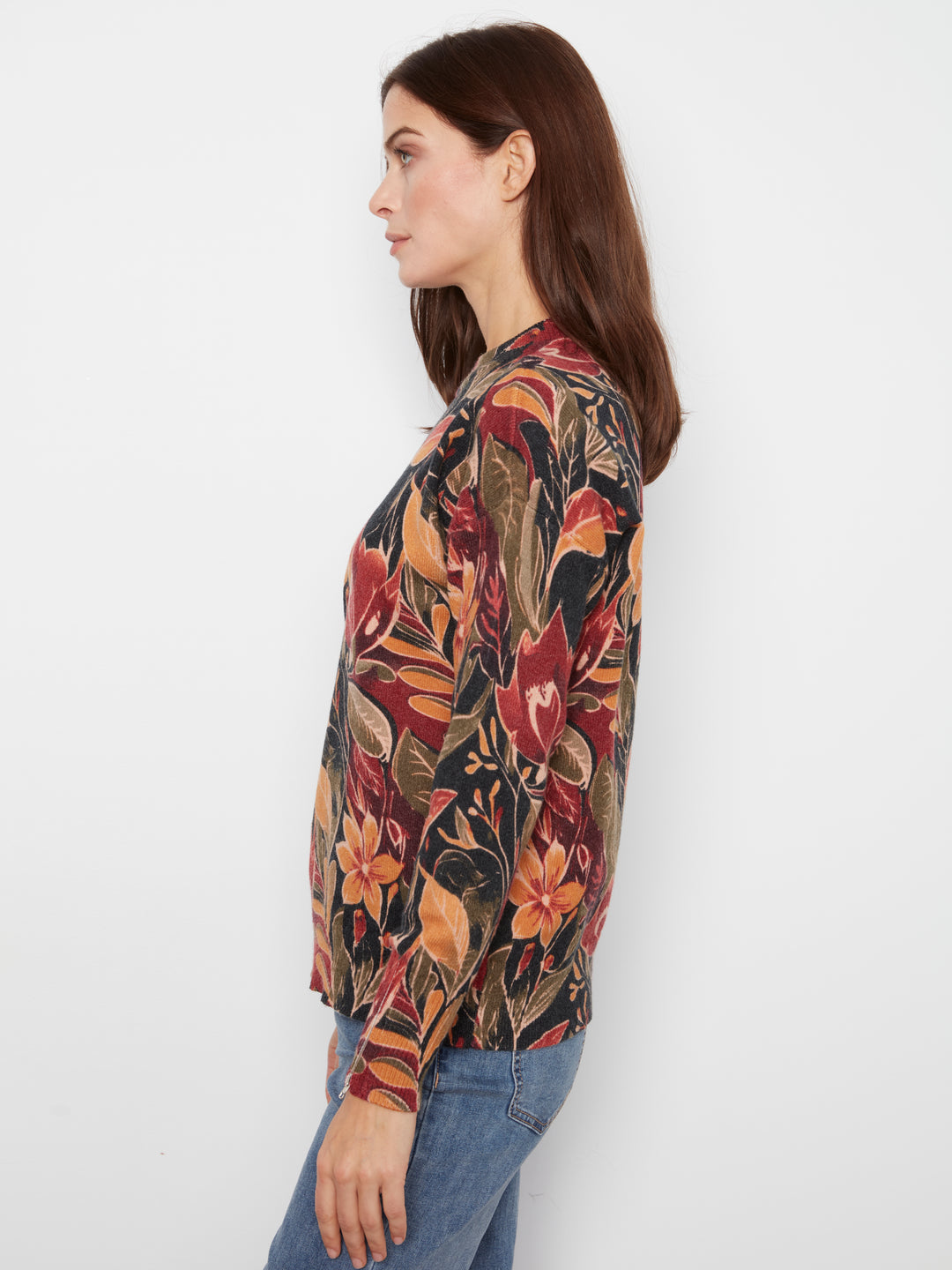 A woman shown in side profile wearing a long-sleeve button-up blouse with a dense floral and botanical print in red, orange, brown, and olive tones, paired with blue jeans against a plain white studio background.