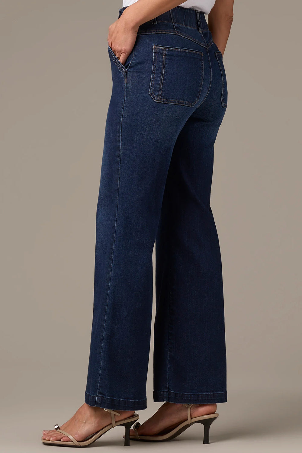 A person wearing dark blue wide-leg denim jeans with a white top and beige heeled sandals, shown from the waist down against a plain beige studio background.