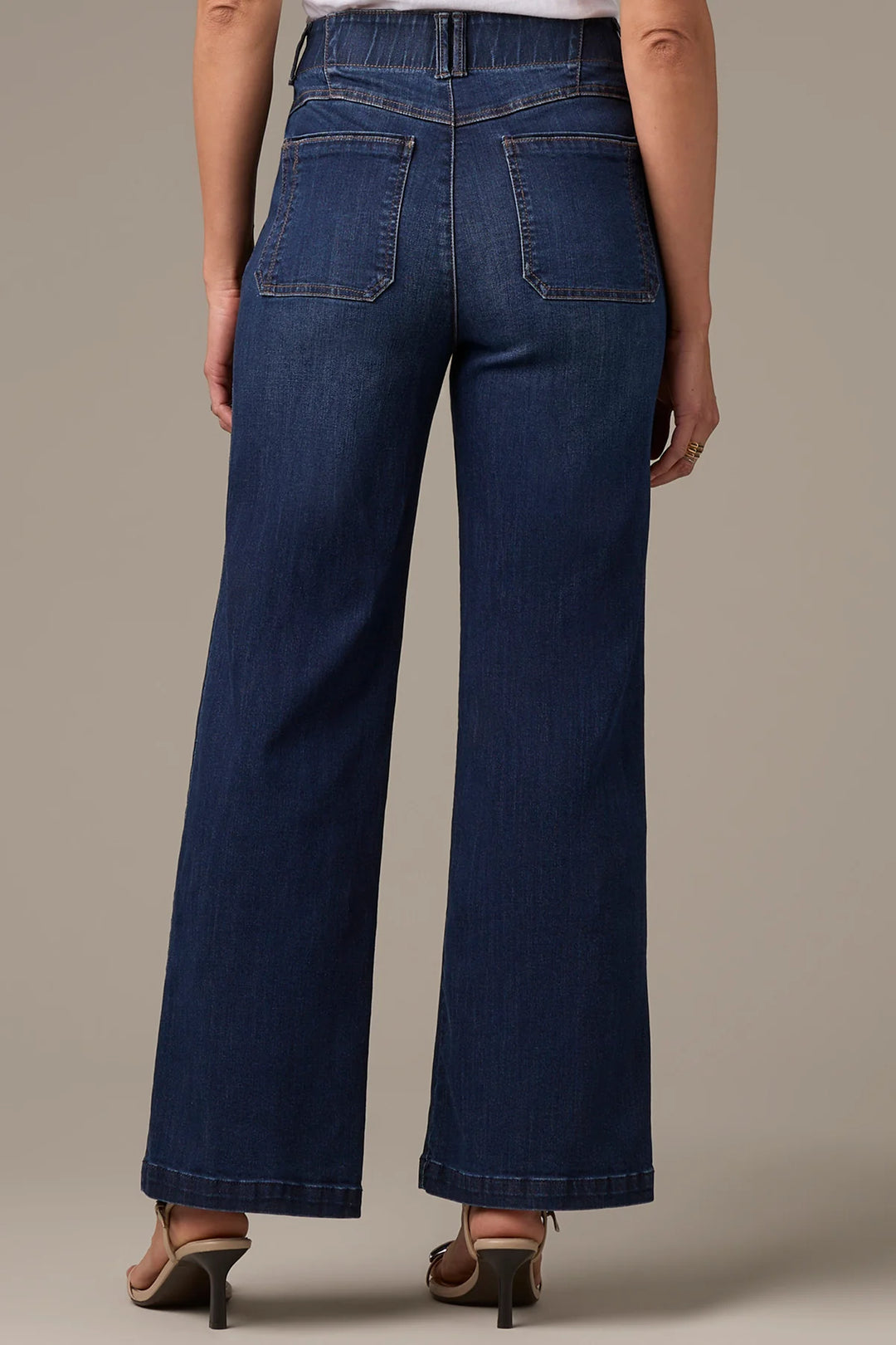 Rear view of a model wearing dark blue wide-leg denim jeans with a high waist and back pockets, paired with brown heeled sandals against a solid beige studio background.