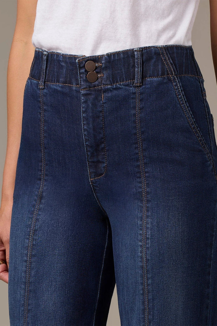 Close-up of a person wearing dark blue denim jeans with a white top, showing the waistband, double button closure, and front pockets.