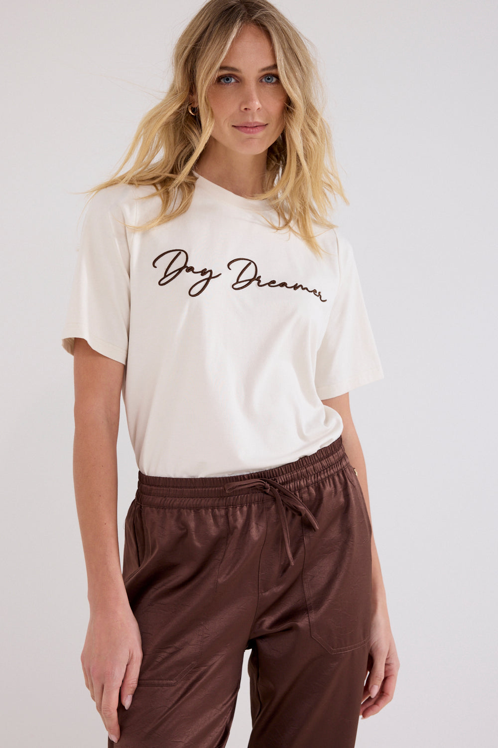 A woman wearing a cream-colored short-sleeve t-shirt with 'Day Dreamer' written in black cursive across the chest, paired with brown satin drawstring pants, against a light gray studio background.