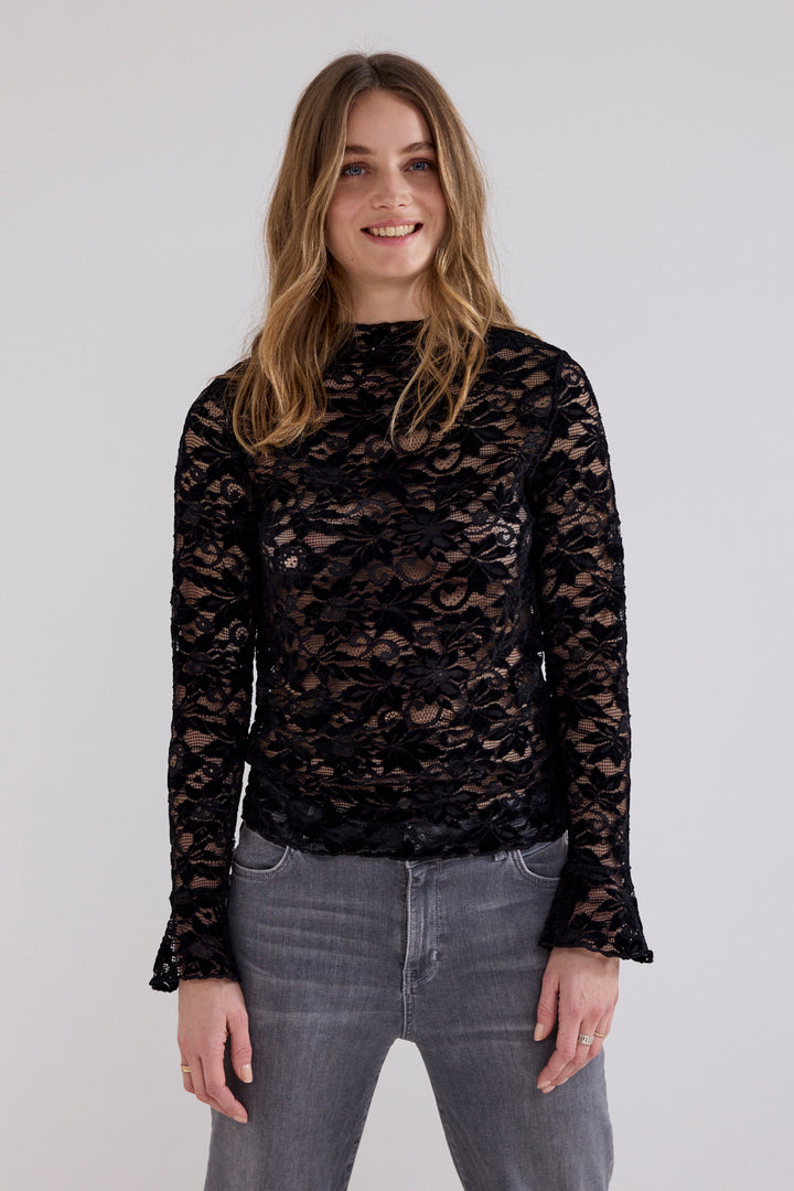 A woman wearing a black lace long-sleeve top and gray jeans, standing against a plain light gray background.