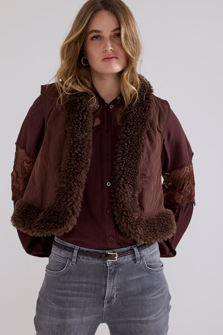 A woman wearing a brown quilted jacket with a brown faux-shearling collar and matching brown lace-up sleeves, styled over a dark burgundy button-up shirt and gray jeans against a light gray studio background.