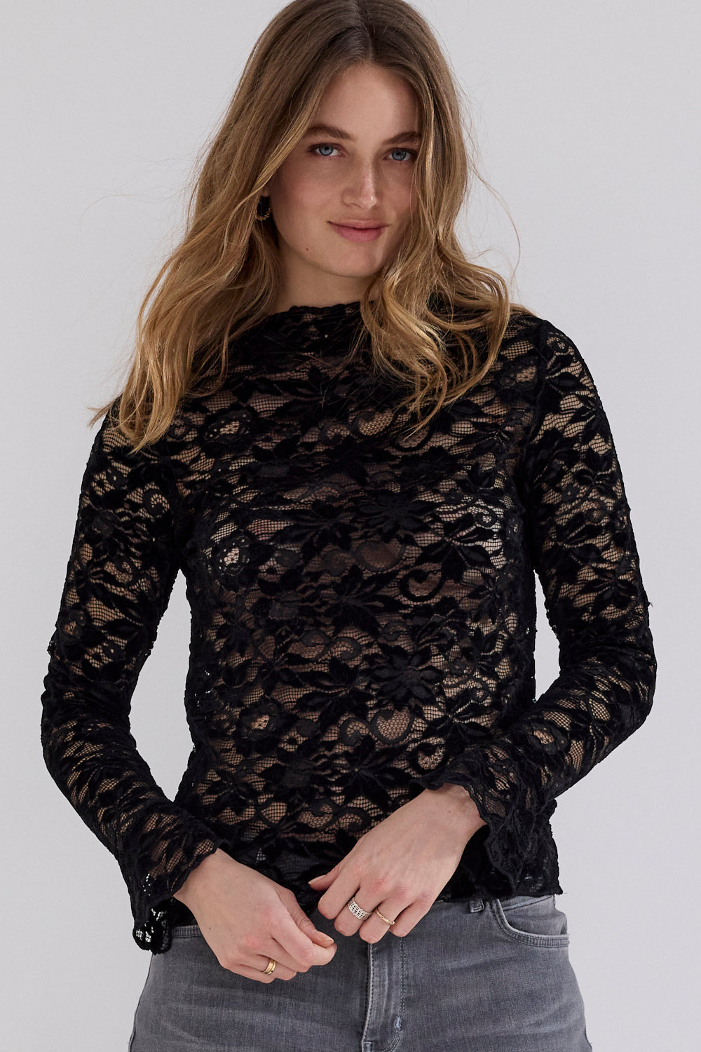 A woman wearing a black floral lace long-sleeve mock neck top paired with gray jeans, photographed against a solid light gray studio background.