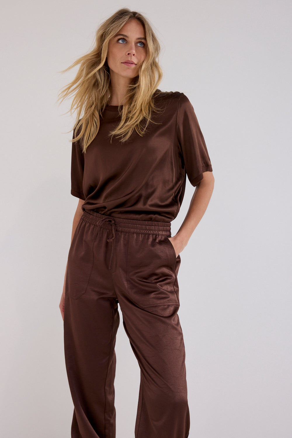 A woman modeling a matching brown satin short-sleeve top and wide-leg pants set against a plain light gray studio background.