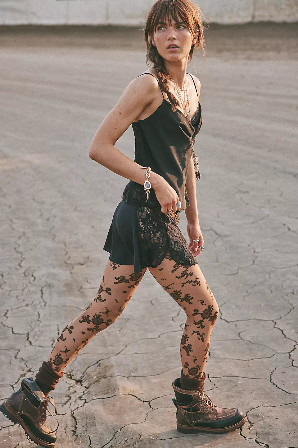 A woman walking outdoors on cracked pavement wearing a black sleeveless top, black lace skirt, sheer floral-patterned tights, and dark lace-up boots.