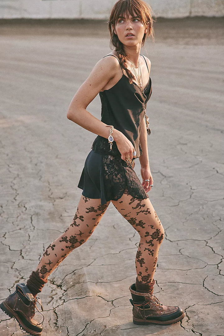 A woman walking outdoors on cracked pavement wearing a black sleeveless top, black lace skirt, sheer floral-patterned tights, and dark lace-up boots.