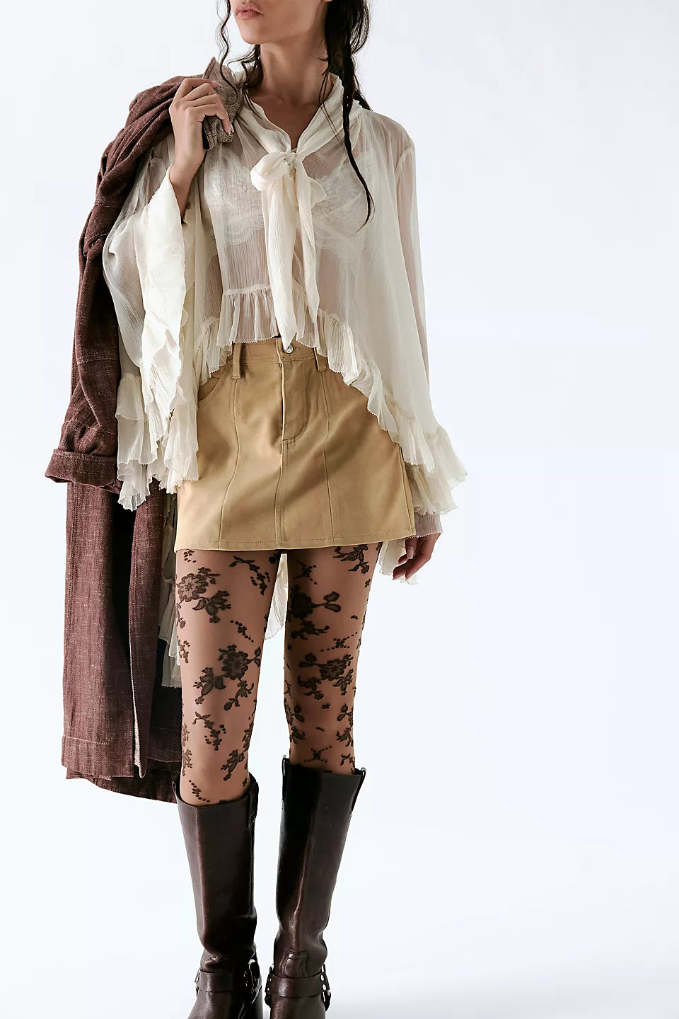 A model wearing a cream ruffled blouse with a tie-neck, a tan mini skirt, floral tights, and dark brown knee-high boots, while holding a brown jacket against a plain white studio background.