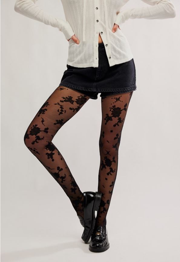 A model wearing sheer black floral lace tights, a black denim mini skirt, a white ribbed cardigan, and black leather shoes, posed against a light gray studio background.