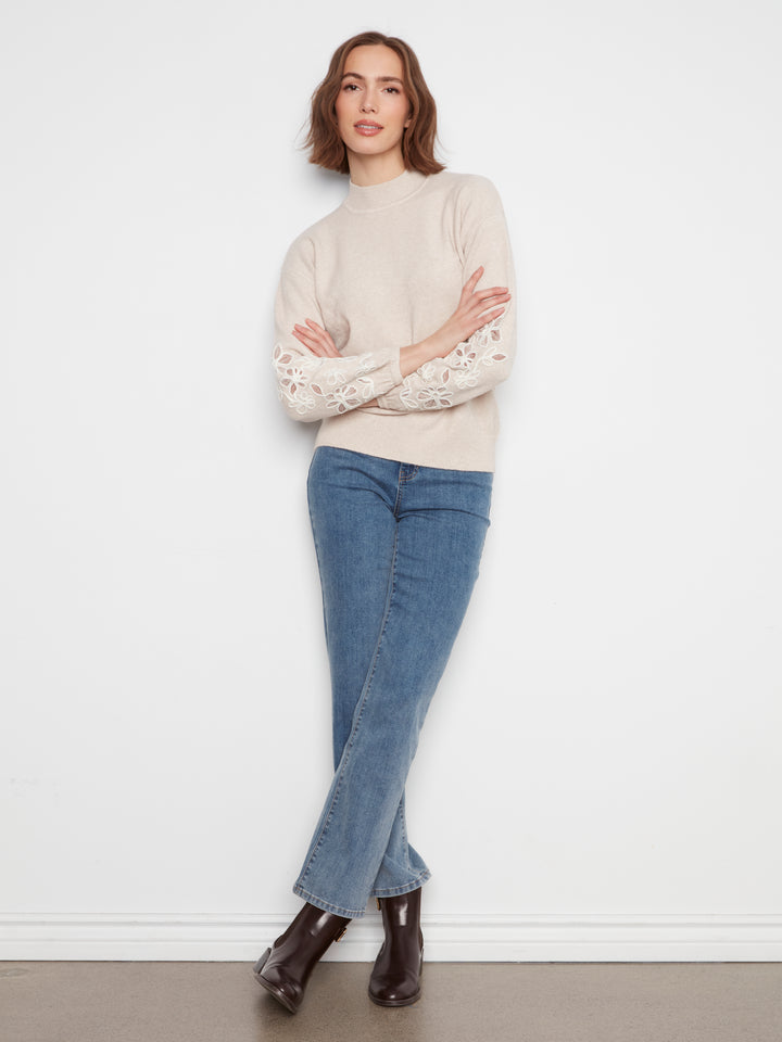 A woman stands against a white wall wearing a cream mock-neck knit sweater with lace sleeves, blue jeans, and dark brown ankle boots.