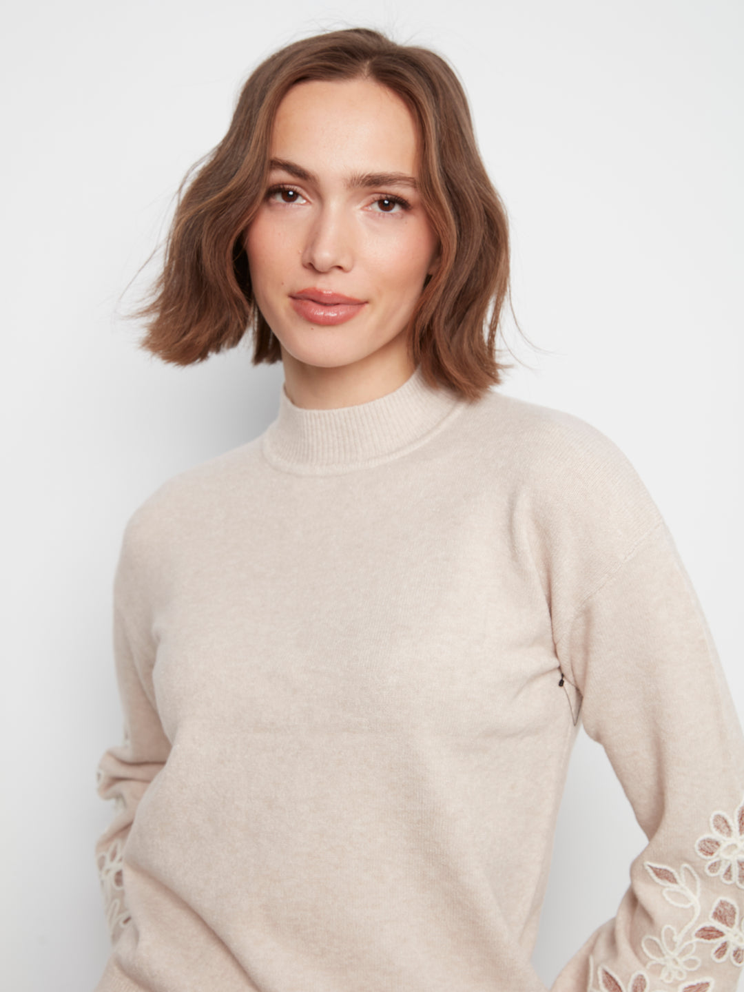 A woman with short brown hair wearing a light beige mock-neck knit sweater with floral lace-up detailing on the sleeves, photographed against a plain light gray background.