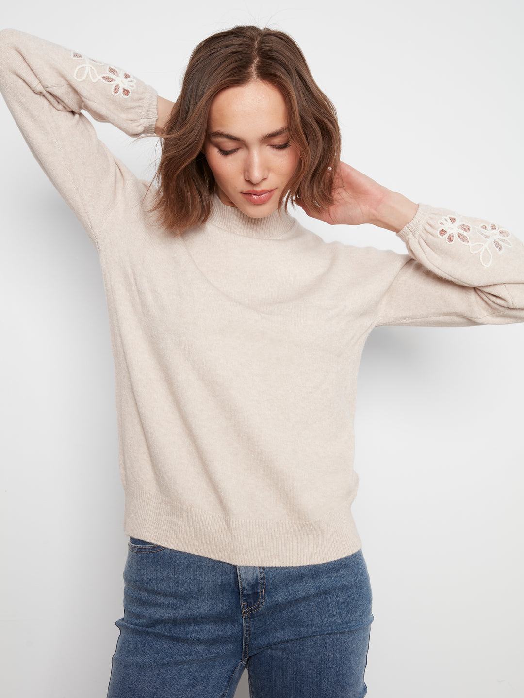 A woman wearing a light beige knit mock-neck sweater with eyelet lace details on the sleeves, paired with blue jeans, against a plain light gray background.