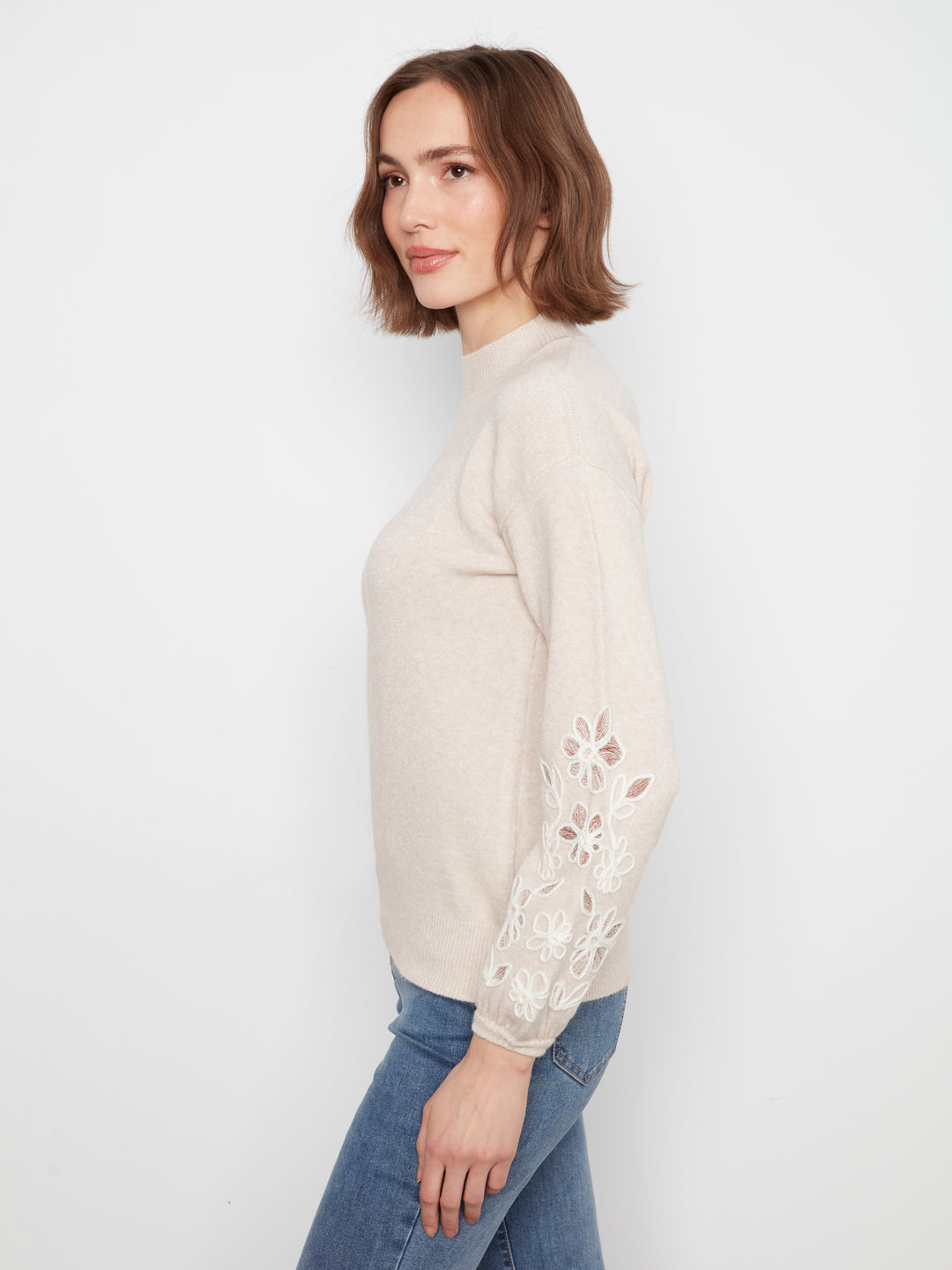 A woman wearing a light beige mock-neck knit sweater with white floral lace embroidery on the sleeves, paired with blue jeans, photographed against a plain white studio background.