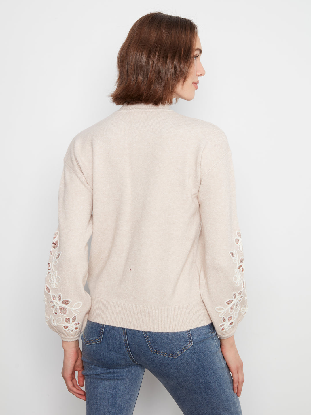 A woman shown from the back wearing a cream-colored knit sweater with intricate floral lace cutout detailing on the sleeves, paired with blue jeans, against a plain white studio background.