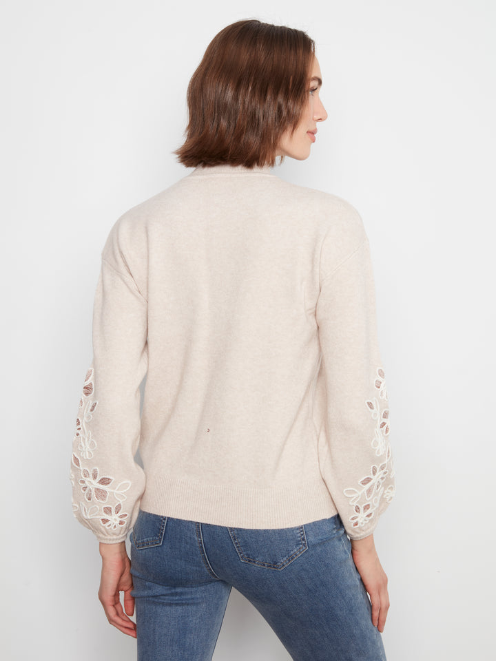 A woman shown from the back wearing a cream-colored knit sweater with intricate floral lace cutout detailing on the sleeves, paired with blue jeans, against a plain white studio background.