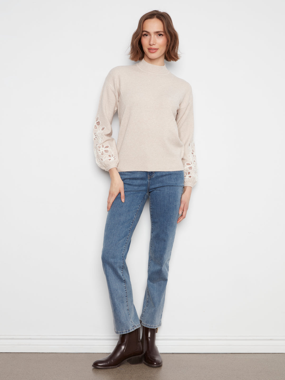 A woman modeling a beige mock-neck knit sweater with lace-up sleeves, paired with blue jeans and dark brown leather ankle boots, standing against a white wall.