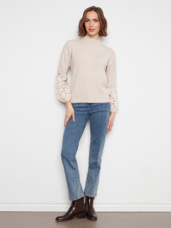 A woman modeling a beige mock-neck knit sweater with lace-up sleeves, paired with blue jeans and dark brown leather ankle boots, standing against a white wall.