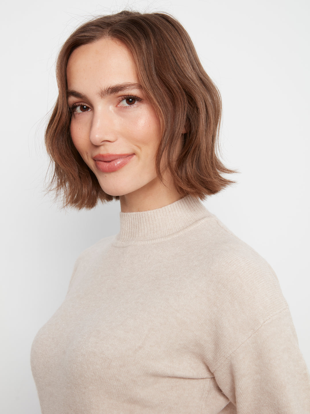 A woman with a short brown bob haircut wearing a light beige knit mock neck sweater, photographed against a plain light gray background.