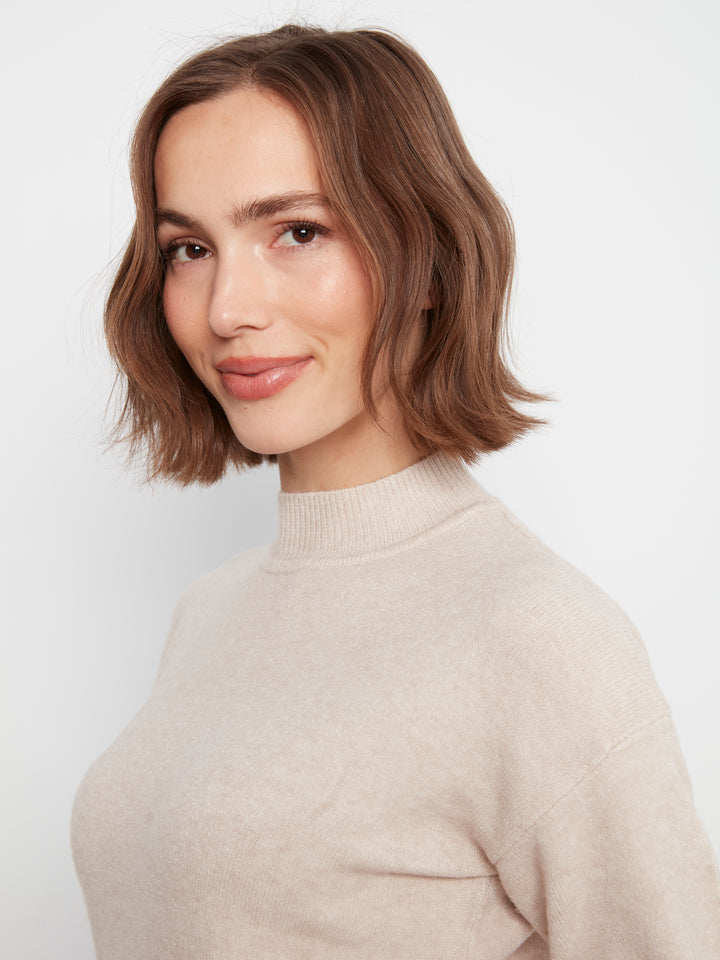 A woman with a short brown bob haircut wearing a light beige knit mock neck sweater, photographed against a plain light gray background.