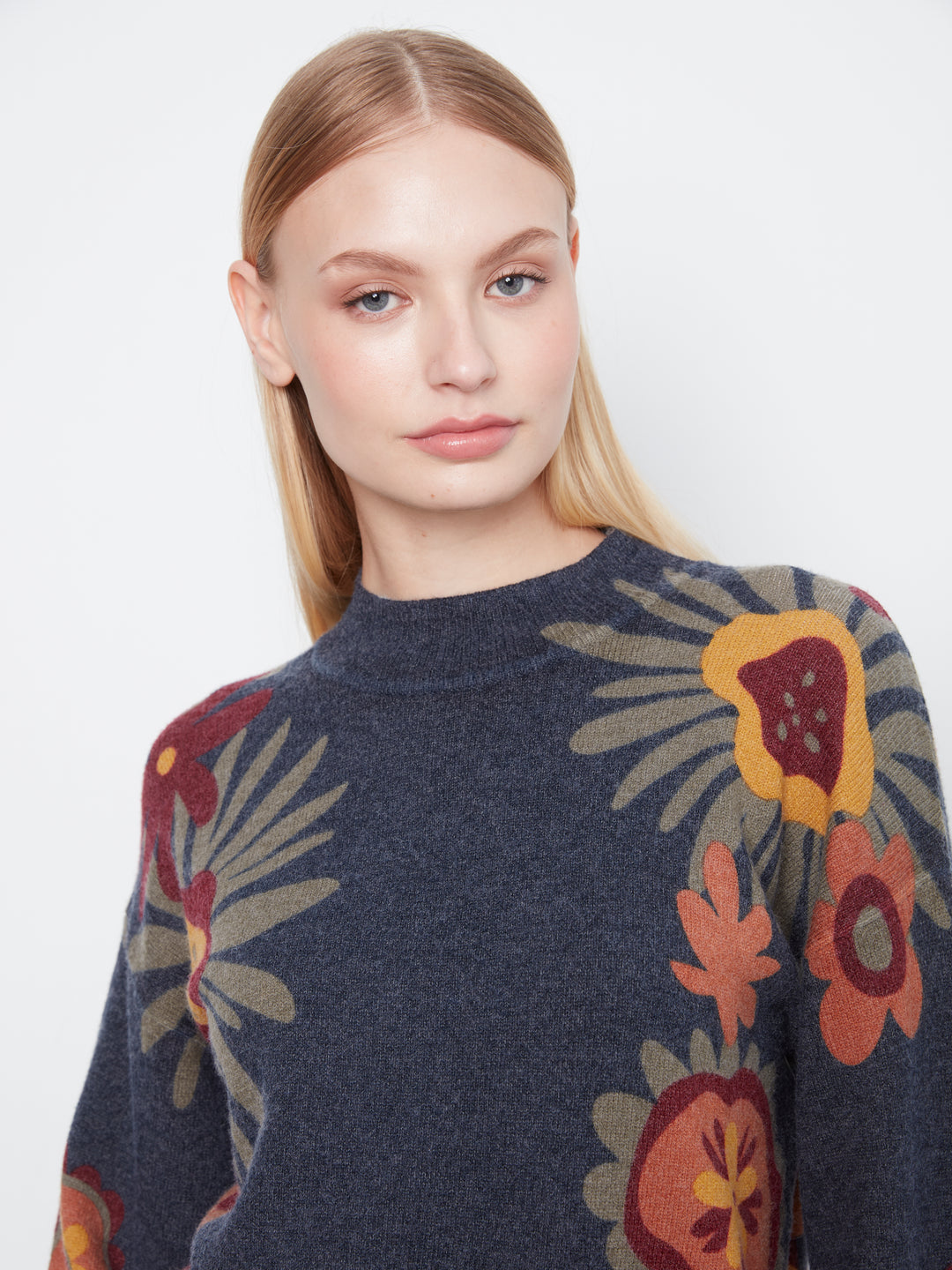A woman wearing a dark navy blue knit sweater with a large, colorful floral pattern in yellow, rust red, and beige, photographed against a plain light gray background.