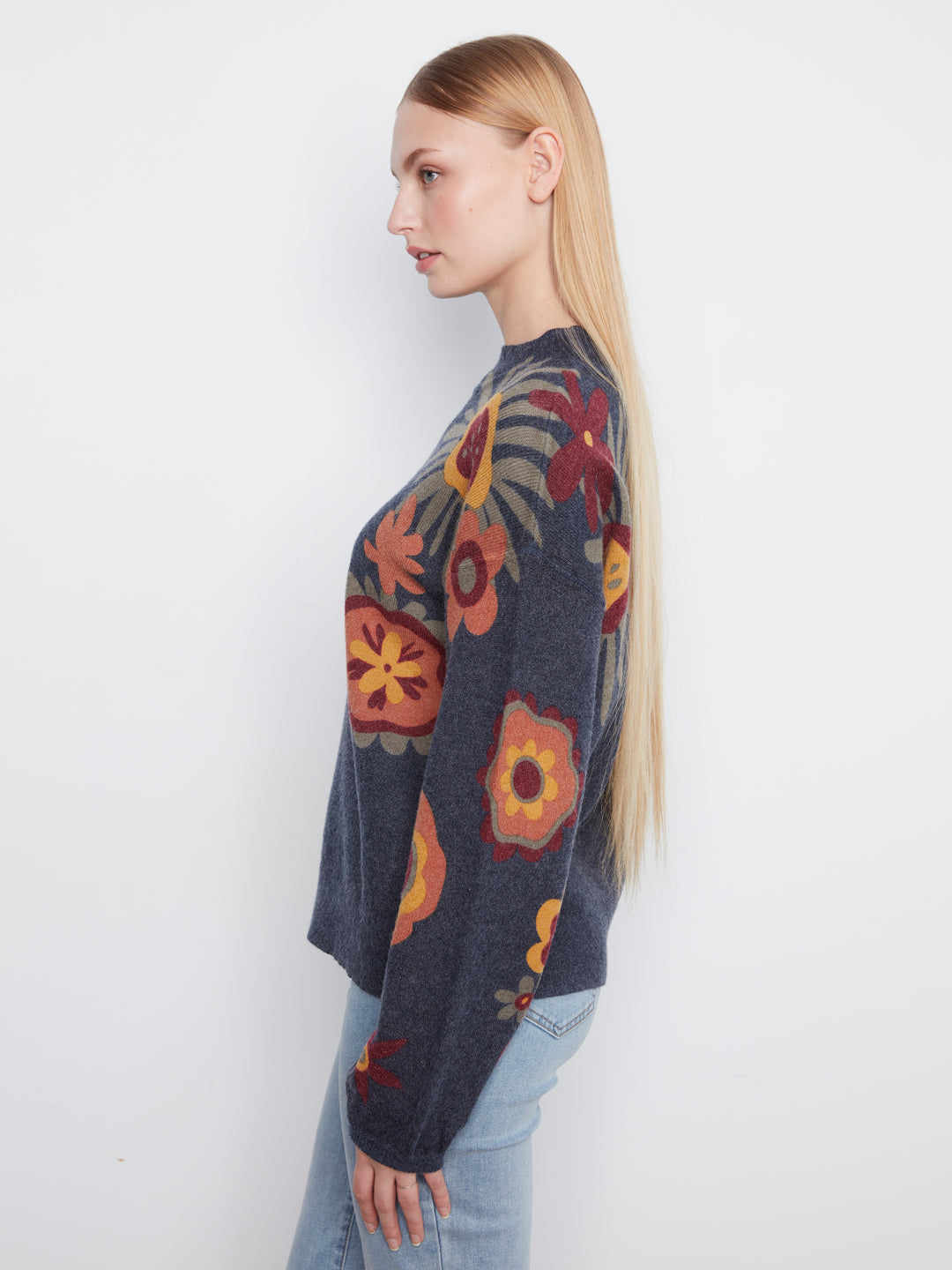 A woman in profile wearing a dark gray knit sweater with a colorful floral pattern in orange, yellow, and red, paired with light blue jeans, against a plain white background.