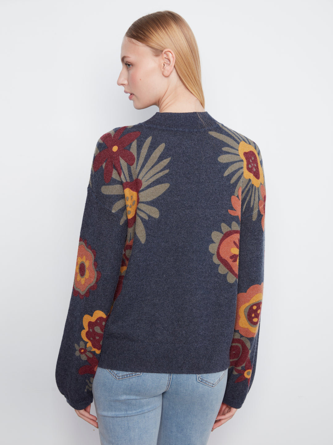 A woman shown from the back wearing a dark navy blue knit sweater with a large floral pattern in red, yellow, and beige, paired with light blue jeans against a plain white background.