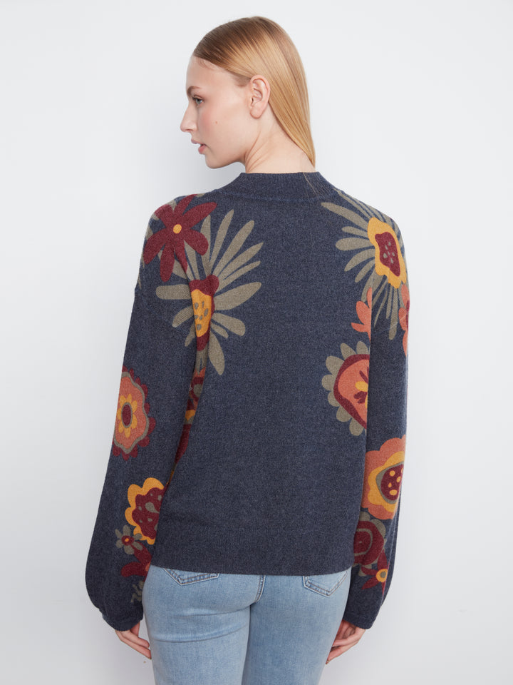 A woman shown from the back wearing a dark navy blue knit sweater with a large floral pattern in red, yellow, and beige, paired with light blue jeans against a plain white background.