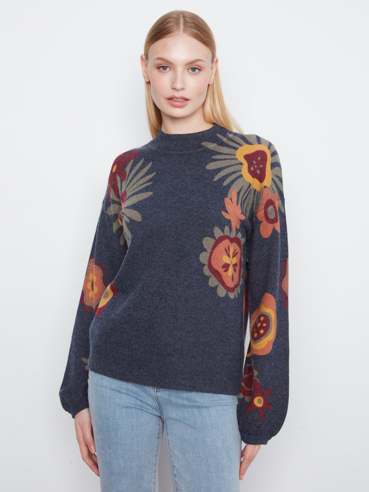A woman modeling a dark navy blue knit sweater with a colorful floral and botanical pattern in red, orange, yellow, and beige, paired with light blue jeans against a plain white background.