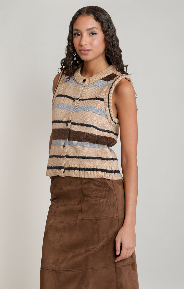 A woman modeling a sleeveless knit vest with horizontal stripes in beige, brown, and light blue, paired with a brown skirt, against a plain light gray studio background.