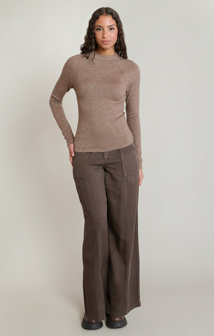 A woman modeling a ribbed long-sleeve mock neck top in taupe brown, paired with dark brown wide-leg trousers, against a solid light gray studio background.