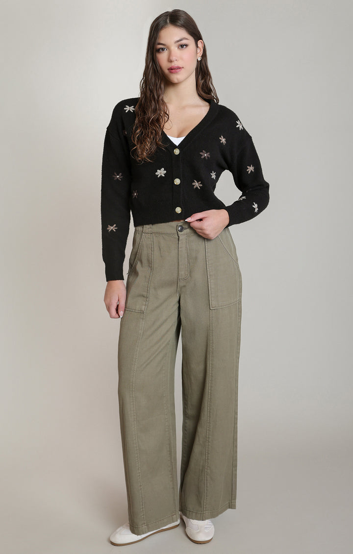 A woman modeling a black long-sleeve knit cardigan with embroidered floral details, paired with high-waisted olive green wide-leg trousers and white sneakers, against a neutral studio background.