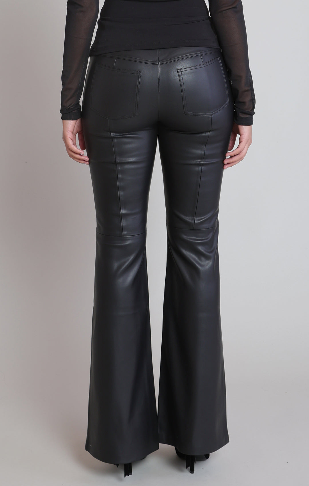 Black faux leather flare pants shown from the back on a model, featuring a smooth leather-like finish and side seam detailing, paired with a black sheer long-sleeve top.