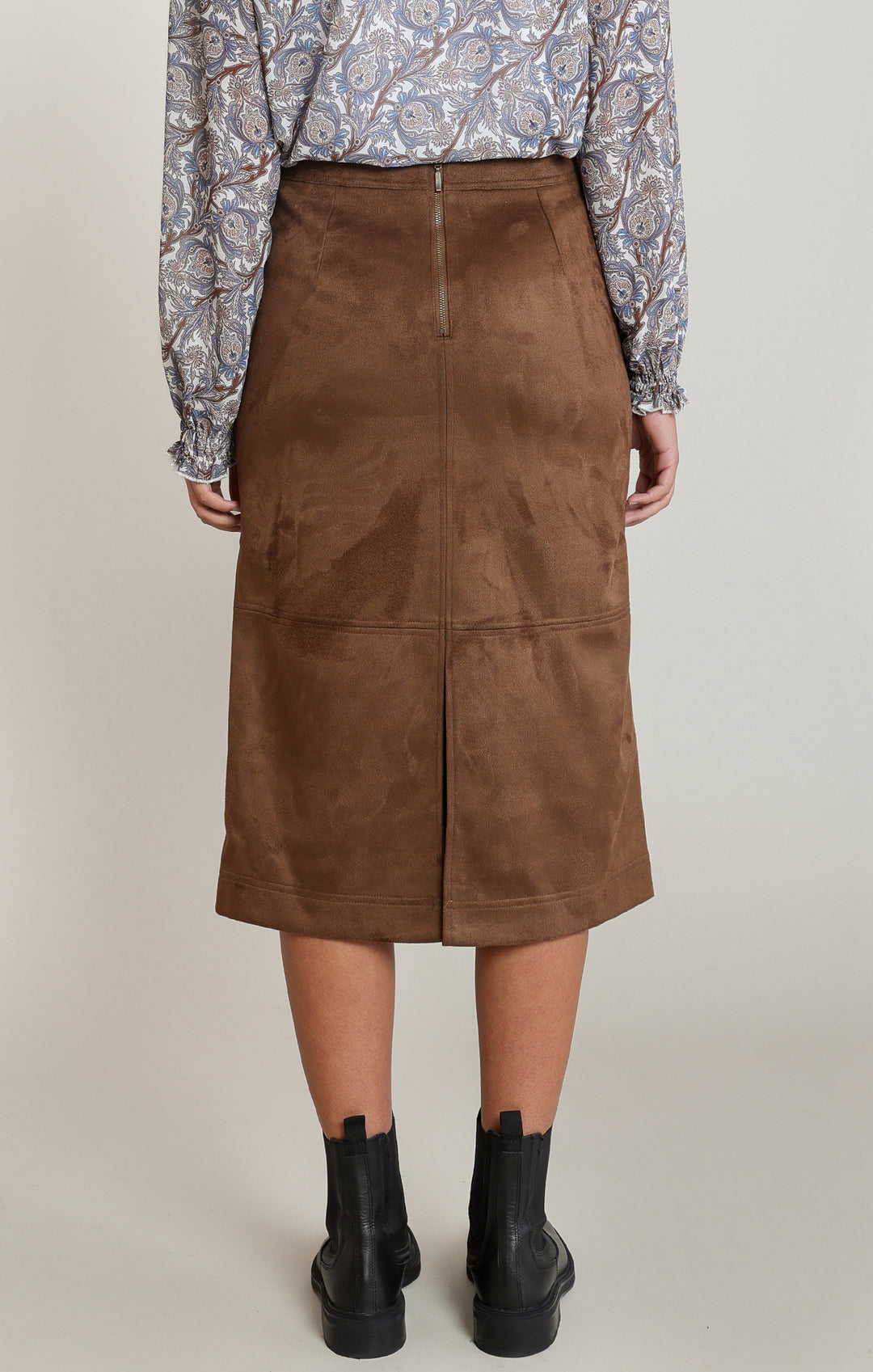 A model is shown from the back wearing a brown suede midi skirt, a patterned long-sleeve top, and black ankle boots against a neutral studio background.