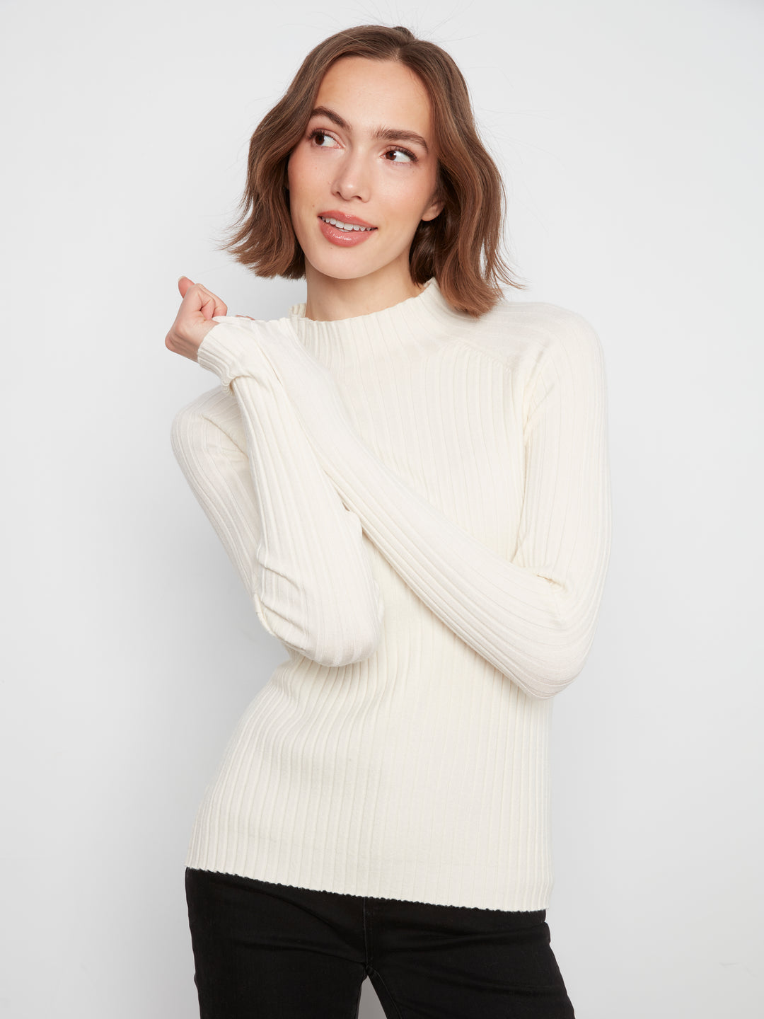 A woman wearing a cream ribbed knit mock neck sweater and black pants, posing against a light gray studio background.