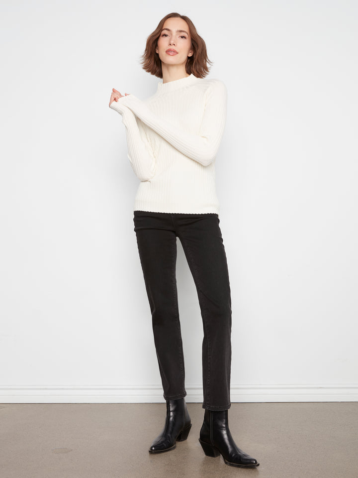 A woman modeling a white mock-neck knit sweater and black skinny pants with black ankle boots, standing against a plain white wall.