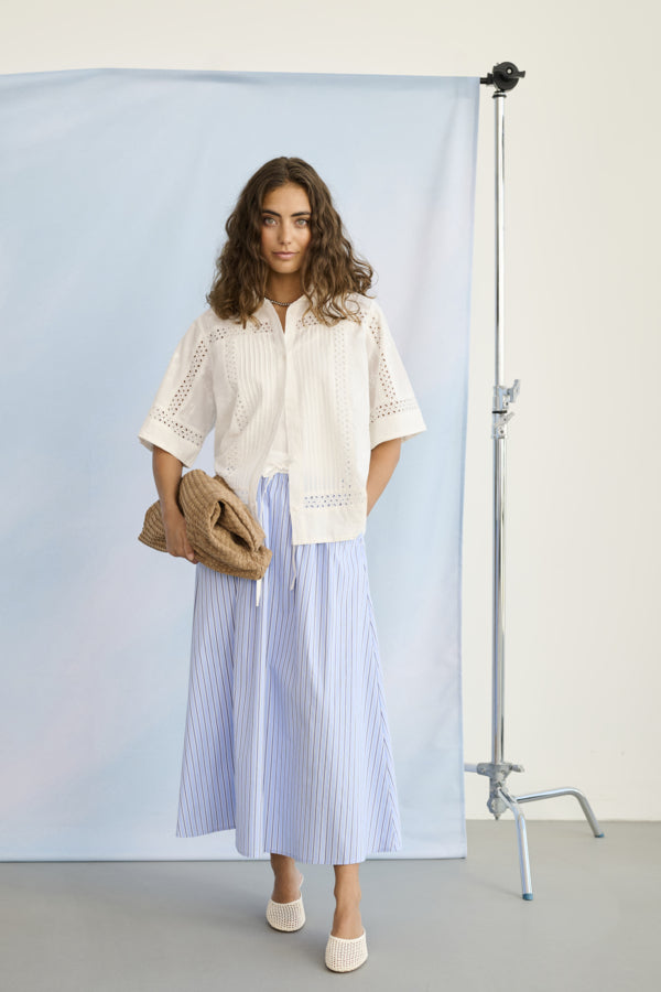 Woman wearing a white blouse and light blue skirt with a beige bag, standing against a light blue backdrop.