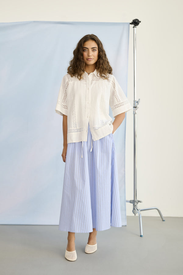 Woman wearing a white blouse and light blue striped pants standing in front of a photography setup with a blue backdrop.