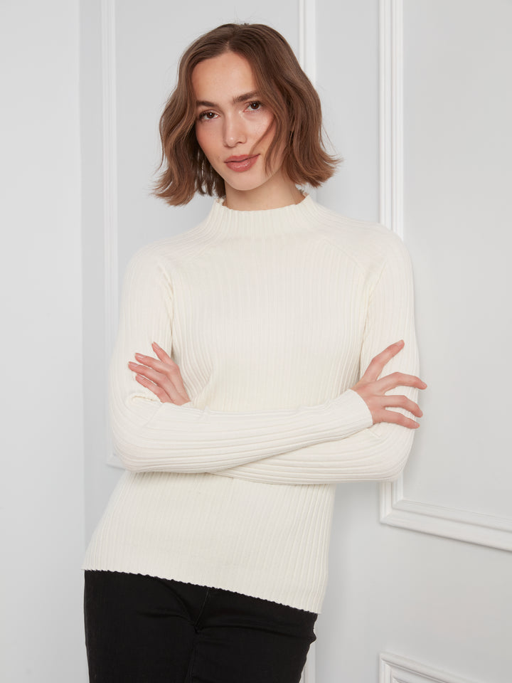 A woman with short brown hair wearing a cream ribbed mock neck sweater and black pants, posing with her arms crossed against a white paneled wall.