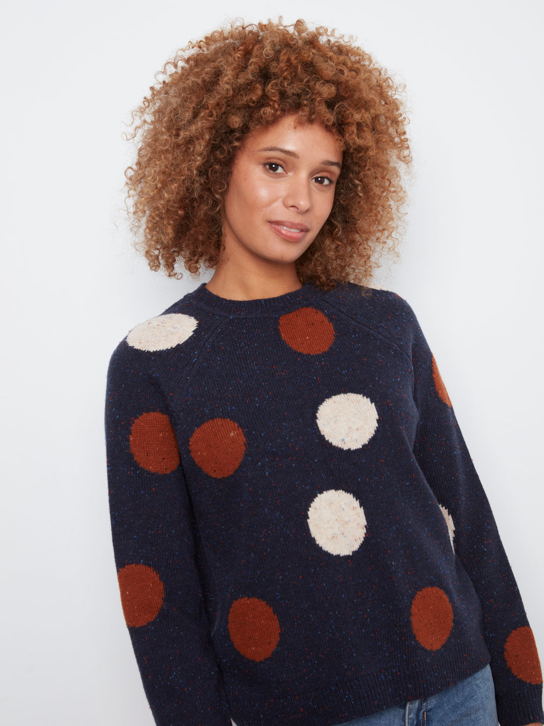 A woman with curly brown hair wearing a navy blue knit sweater with large brown and cream polka dots, paired with blue jeans, photographed against a plain white background.