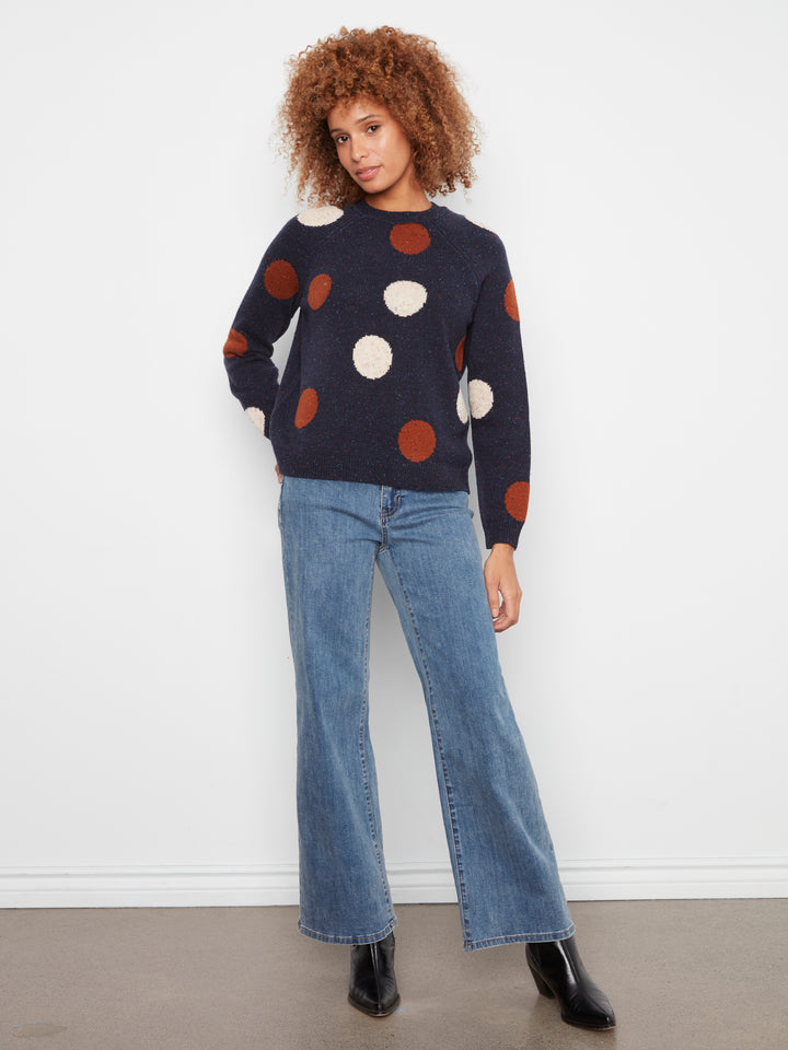 A woman modeling a navy blue knit sweater with large brown and cream polka dots, paired with wide-leg blue jeans and black ankle boots, standing against a plain white wall.