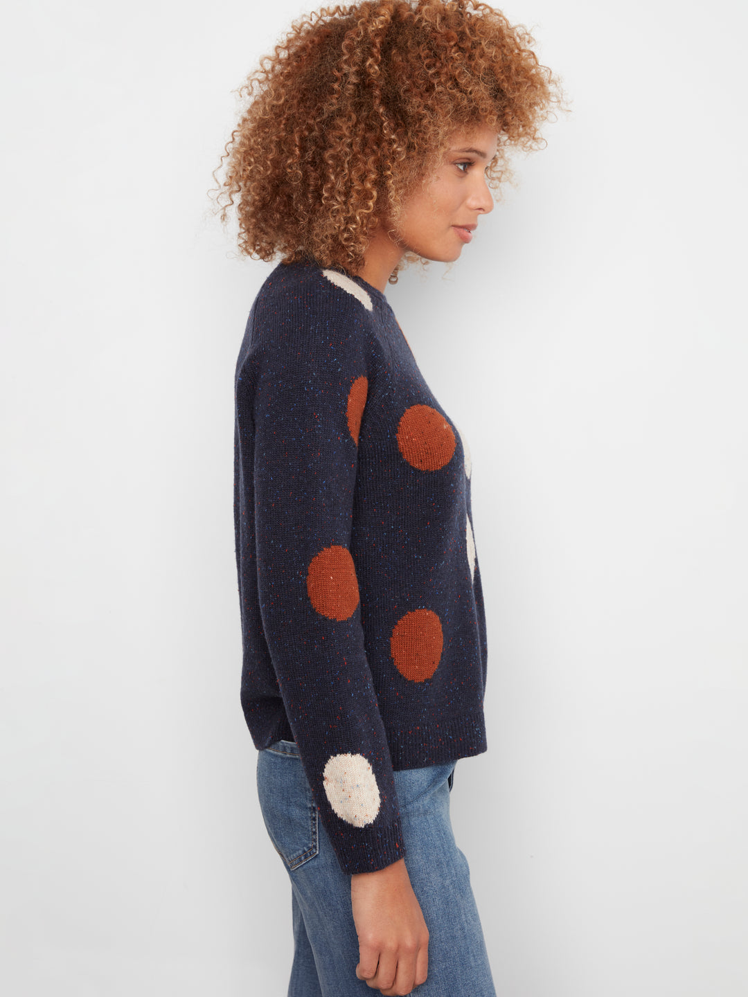 A woman shown in side profile wearing a navy blue knit cardigan with large brown and cream pom-pom style polka dots, paired with blue jeans, against a plain light gray background.
