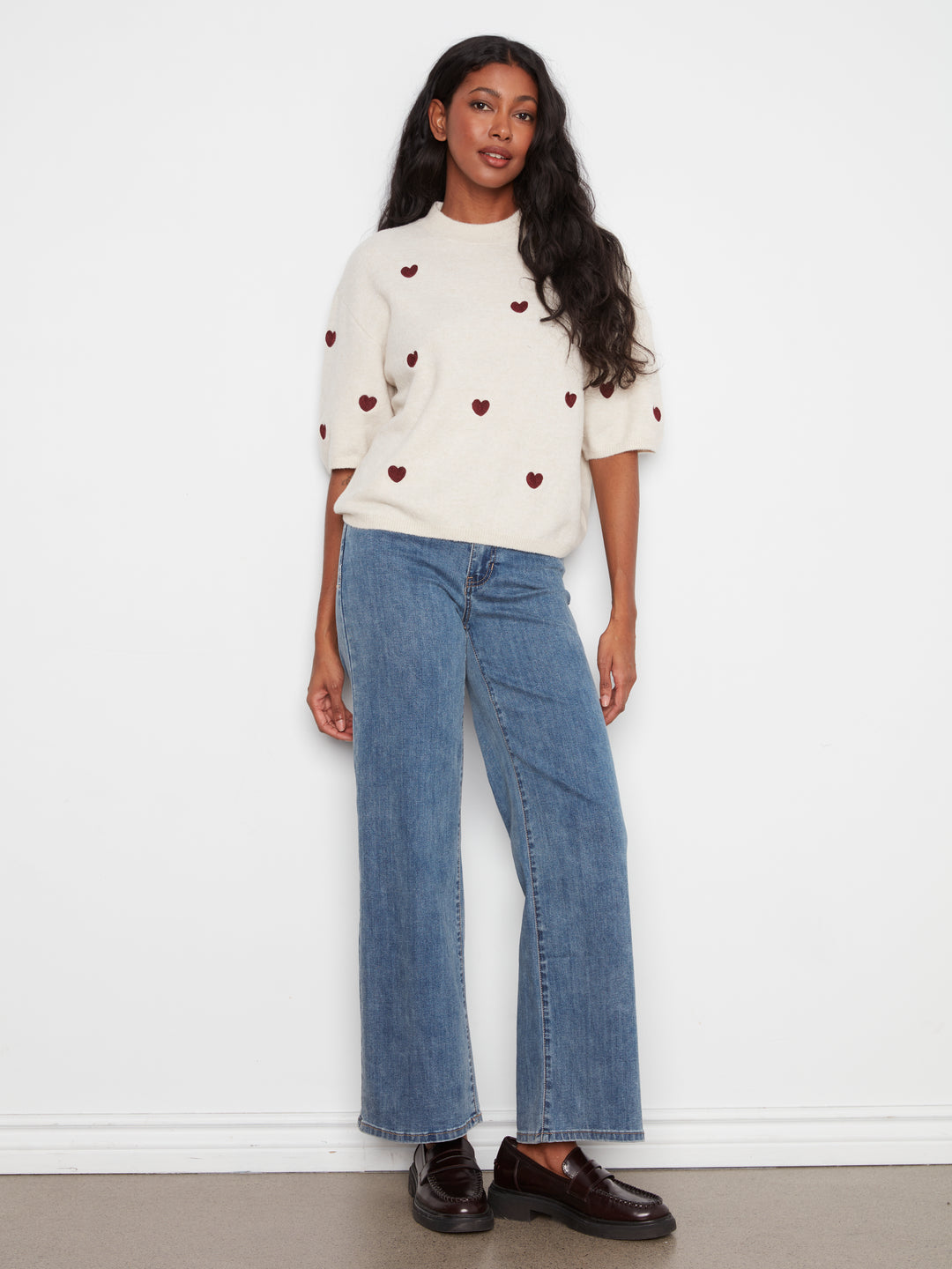 Woman wearing a cream sweater with red heart patterns and blue jeans against a white background