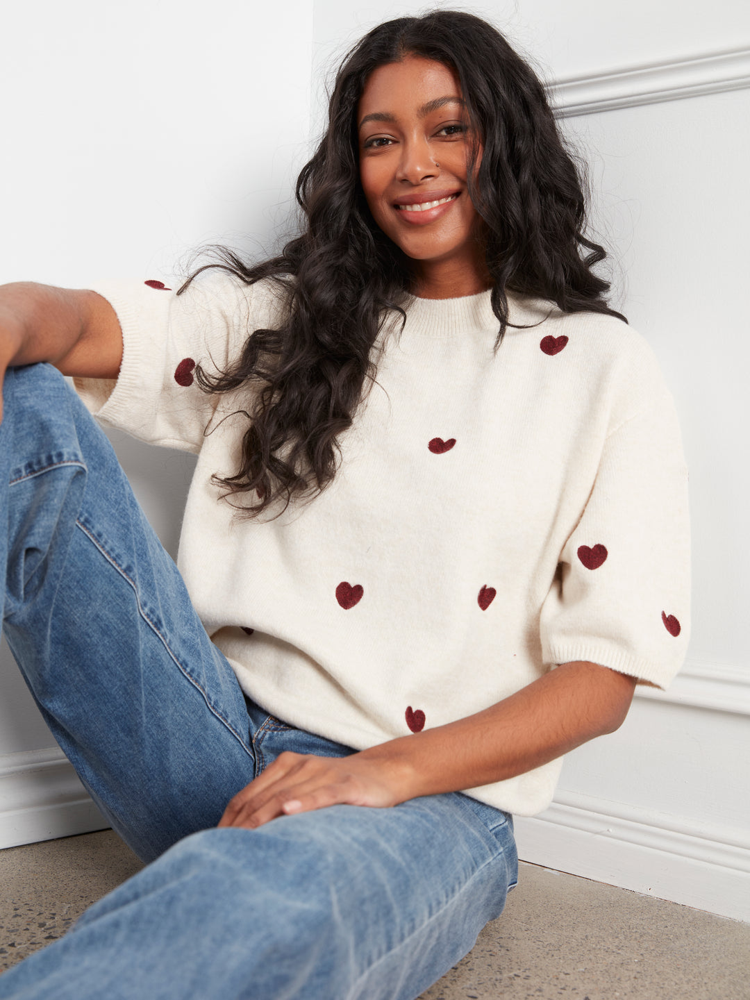 Woman wearing a white sweater with red heart patterns and blue jeans sitting on the floor.