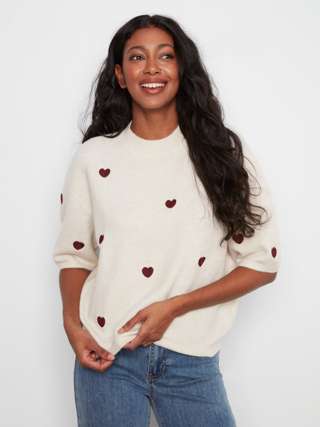 A woman wearing a cream-colored knit sweater with small dark red heart motifs, paired with blue jeans, photographed against a plain light background.