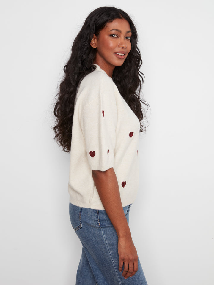 Woman wearing a cream sweater with red heart patterns and blue jeans against a white background